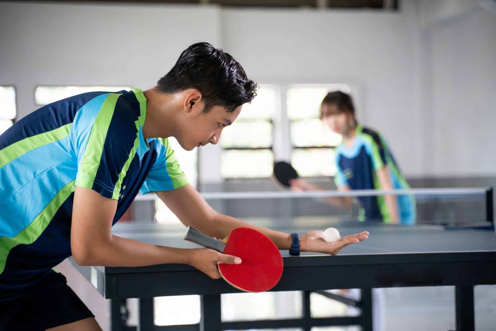 Home Ein junger Tischtennisspieler beim Aufschlag in einer professionellen Indoor-Anlage mit Fokus und Technik.
