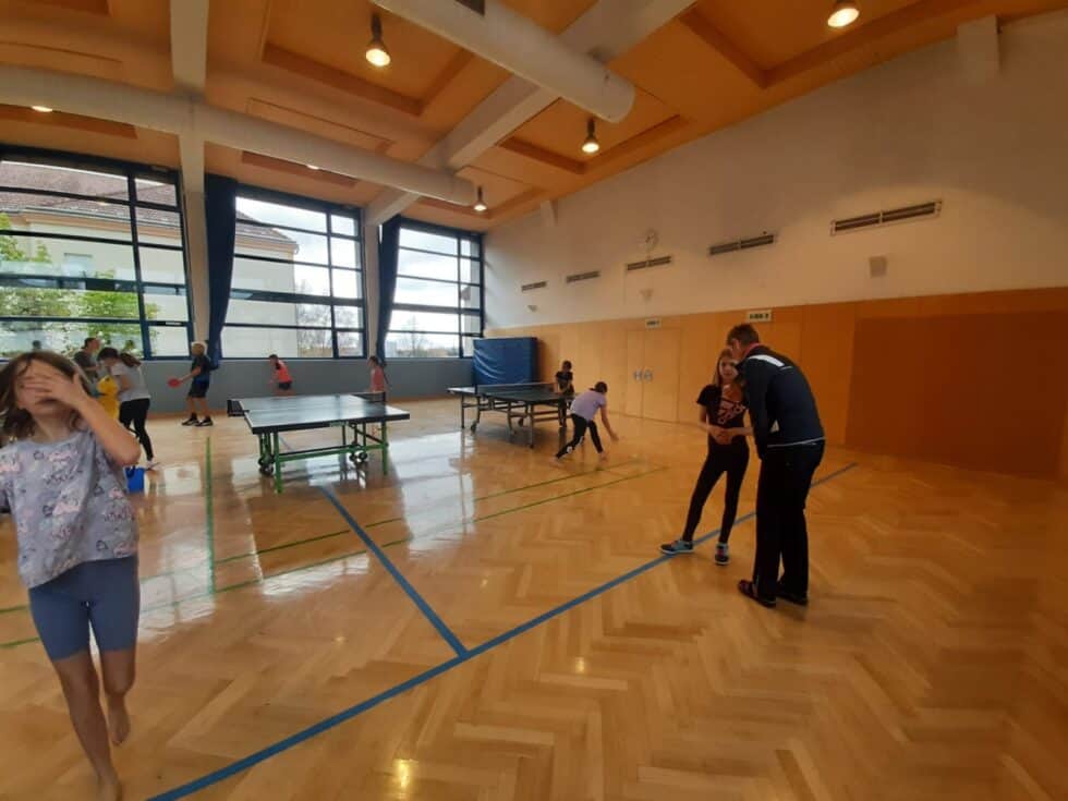 Kinder spielen Tischtennis in einer hellen Turnhalle, betreut von einem Trainer.