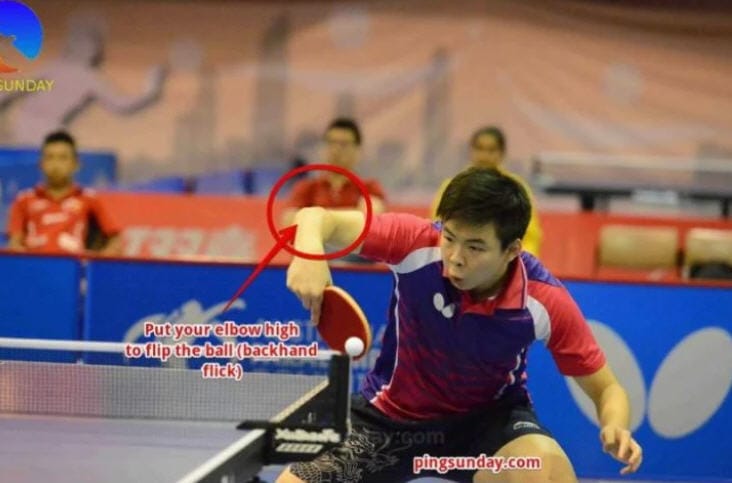 Tischtennisspieler zeigt Backhand Flick Technik mit hohem Ellbogen beim Match in der Halle.