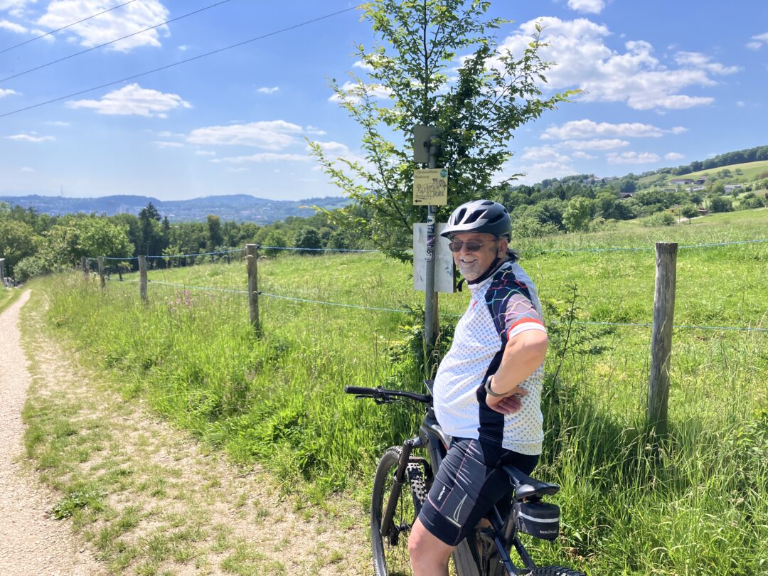 Bilder von unseren Mitgliedern Radfahrer in idyllischer Landschaft auf Schotterweg, umgeben von grünen Hügeln und blauem Himmel.