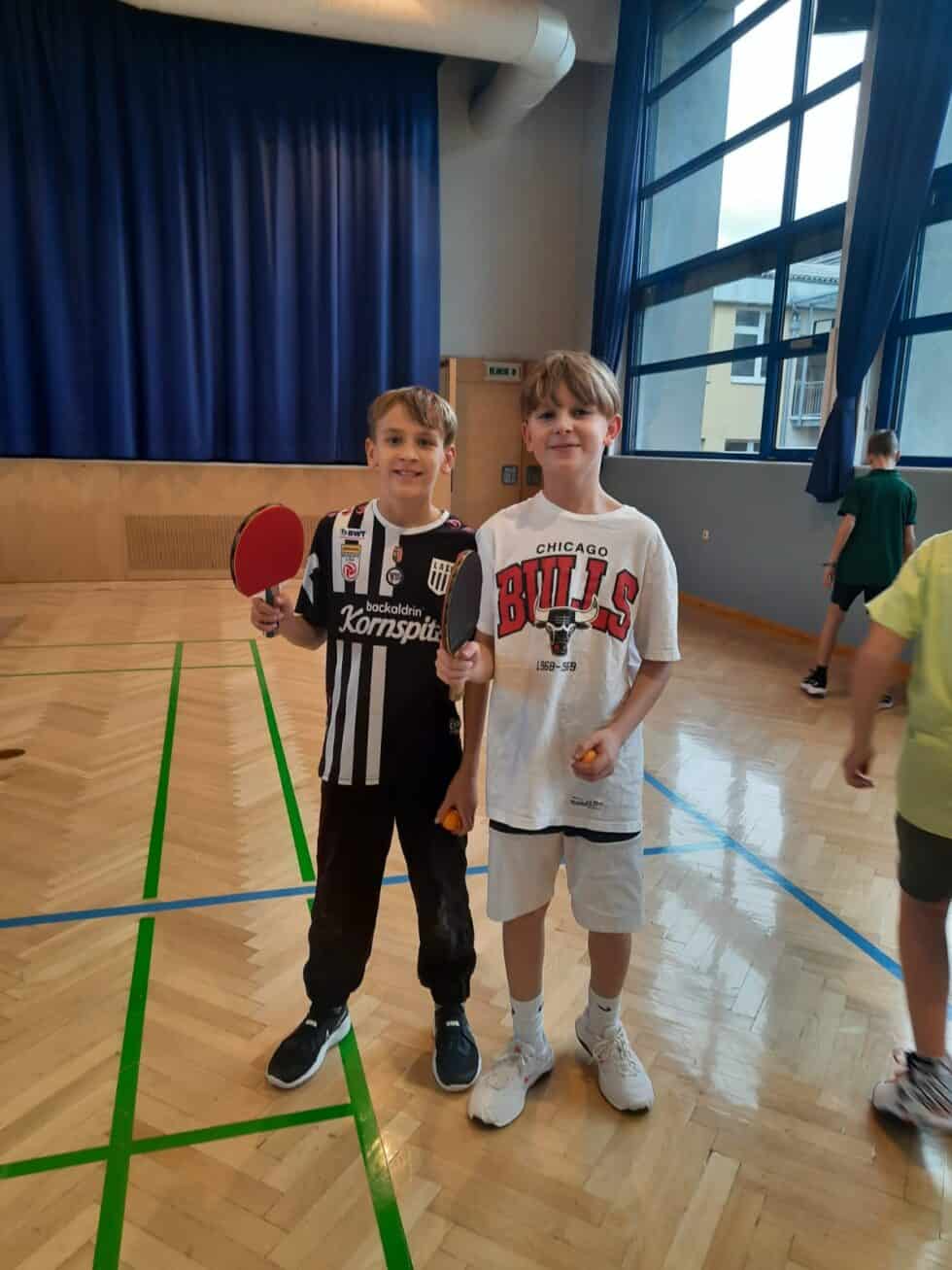 Fröhliche Jungs spielen Tischtennis im lichtdurchfluteten Turnsaal mit Sportkleidung und anderen Kindern.