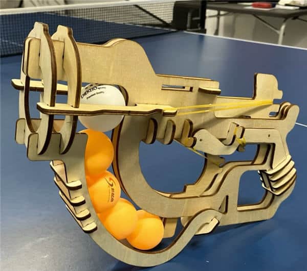 Tischtennis Leonding | Bilder „Tischtennis Leonding“ Holz-Tischtennisballwerfer auf blauem Tisch, mit Gummiband-Antrieb und orangem Ballmagazin.