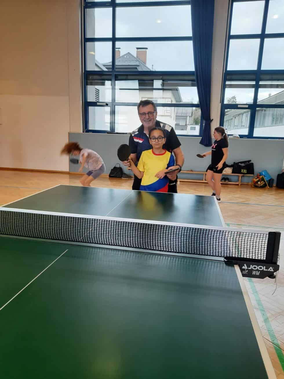 Mann und Mädchen spielen Tischtennis im Sonnenlicht, umgeben von weiteren Kindern im Turnsaal.