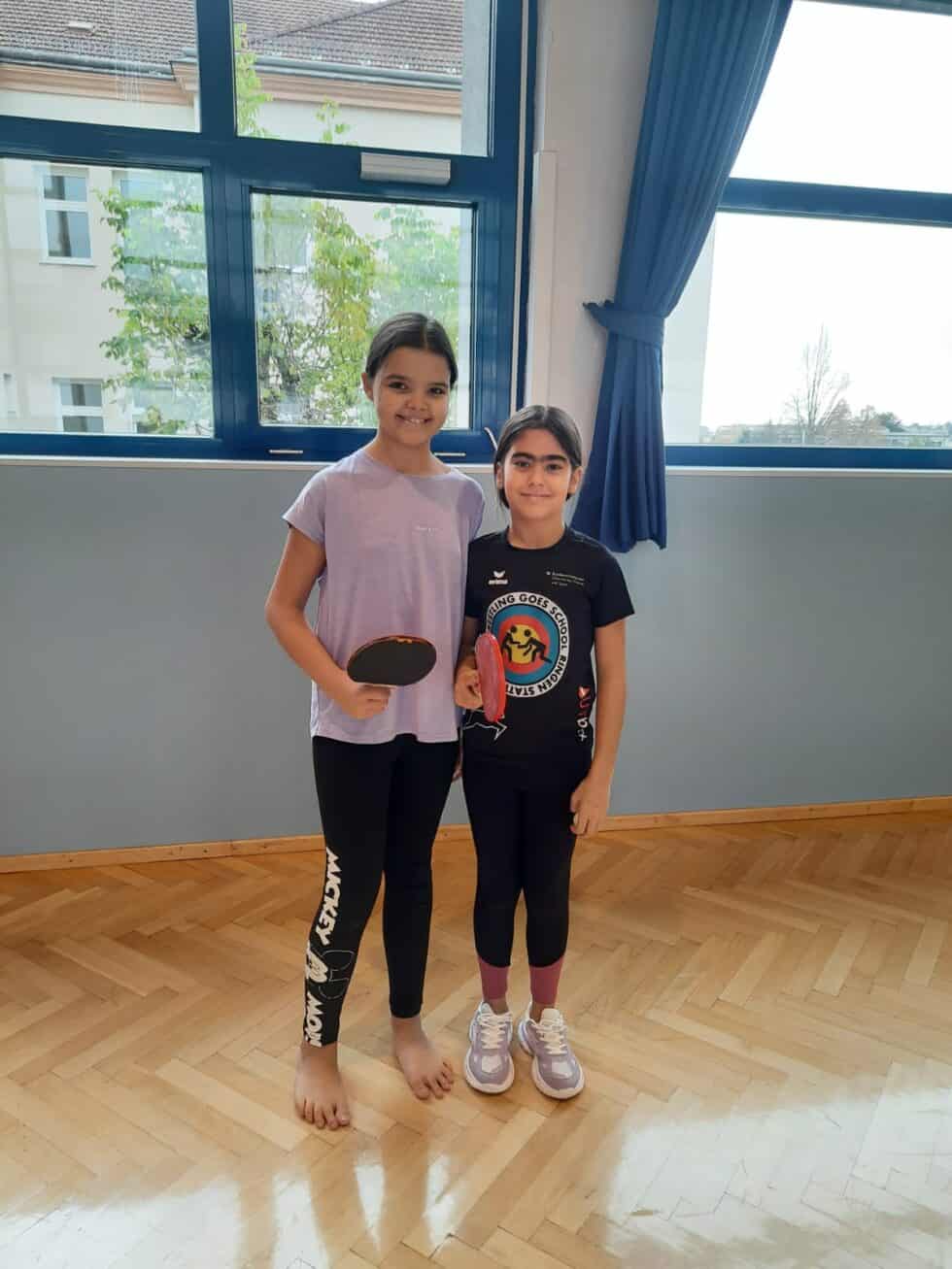 Zwei Mädchen spielen lächelnd Tischtennis in heller Turnhalle mit Holzboden und großen Fenstern.