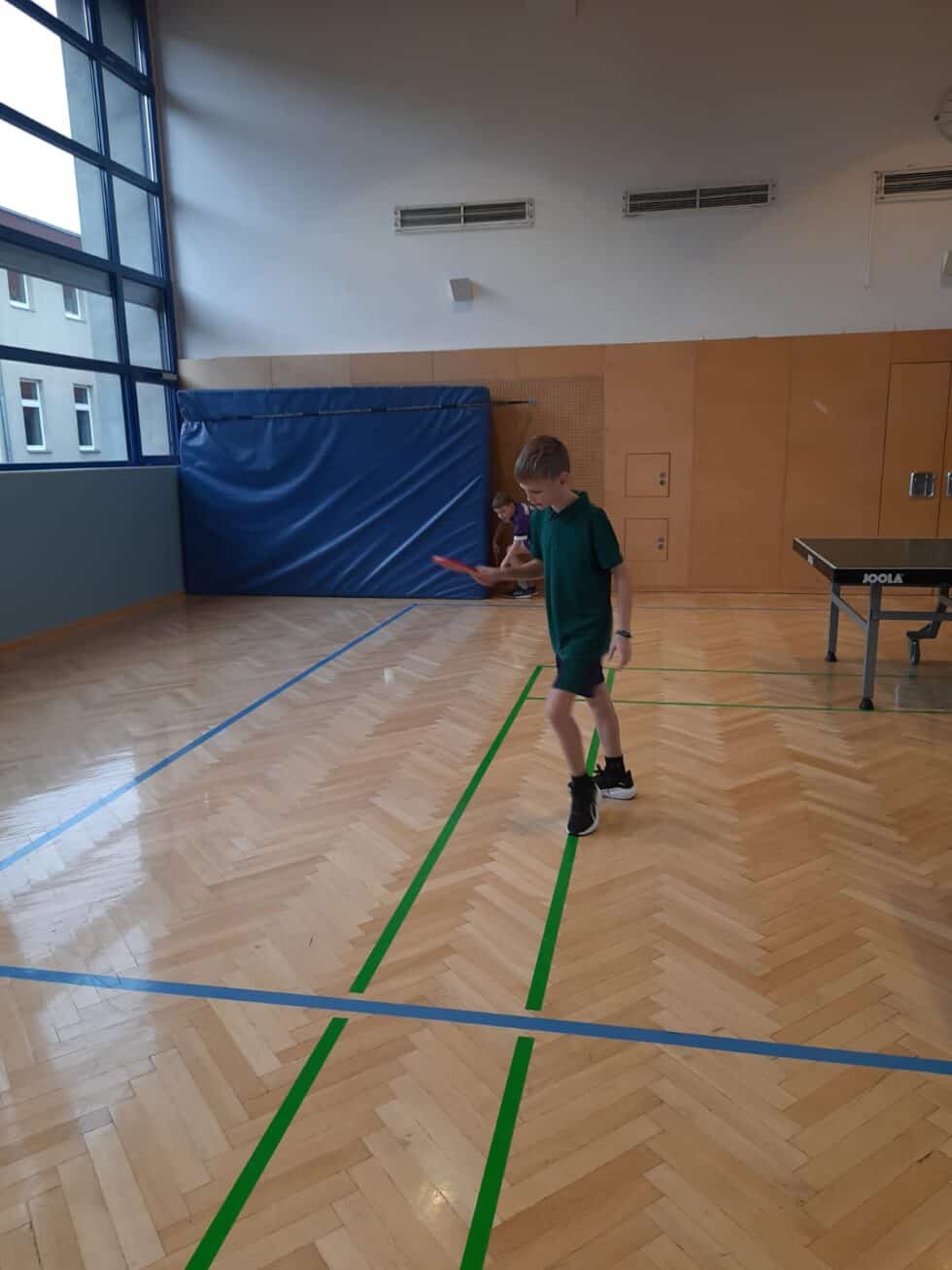 Junge spielt Tischtennis in einem hellen Turnsaal mit JOOLA Tisch und natürlichem Licht.
