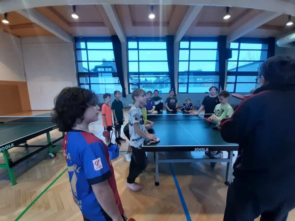 Kinder spielen Tischtennis in heller Halle; Betreuer gibt Anweisungen; Atmosphäre aktiv und teamorientiert.