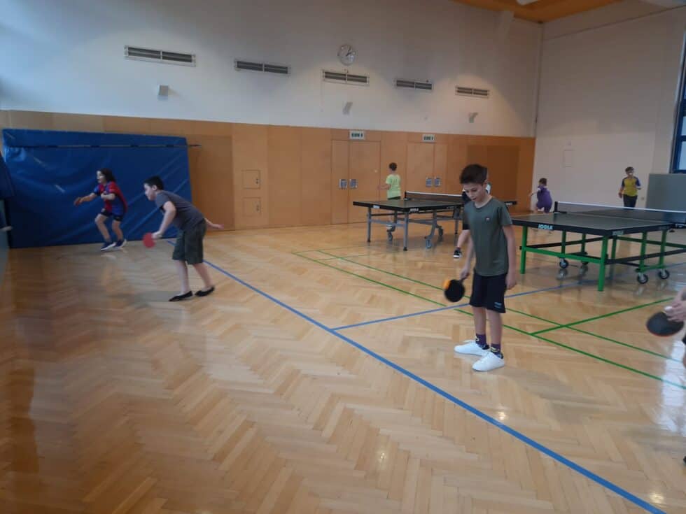 Kinder spielen Tischtennis in heller Turnhalle mit grünen Tischen, Holzfußboden und natürlichem Licht.