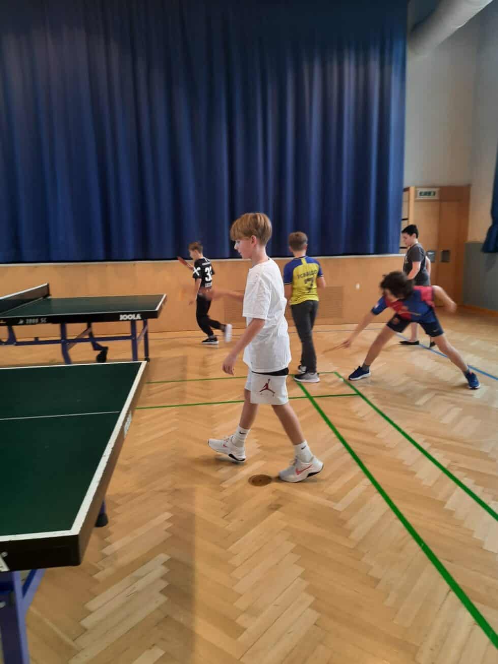 Kinder beim Tischtennis in moderner Turnhalle mit blauem Vorhang und Holzfußboden.