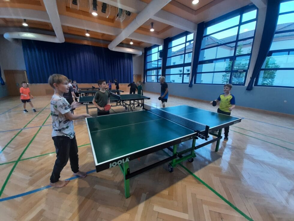 Kinder spielen Tischtennis in einem hellen Turnsaal mit natürlichem Licht und lebhafter Atmosphäre.