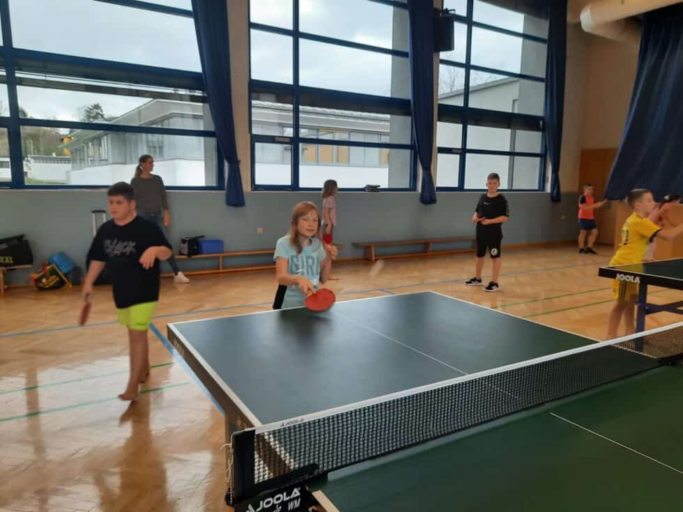 Kinder spielen Tischtennis in hellem Turnsaal, mit Fokus auf Spiel und sozialem Miteinander.