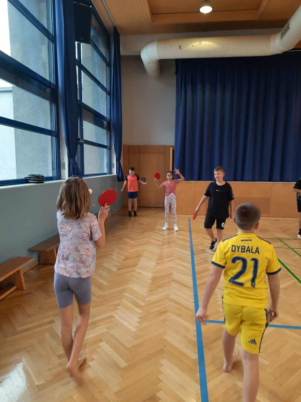 Kinder spielen Tischtennis im sonnendurchfluteten Turnsaal mit blauen Vorhängen und Holzboden.