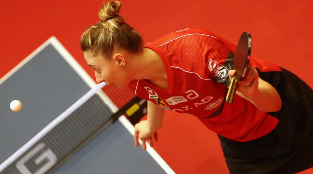 Tischtennisspielerin beim Aufschlag, konzentriert im roten Trikot auf blauem Tisch in Sporthalle.