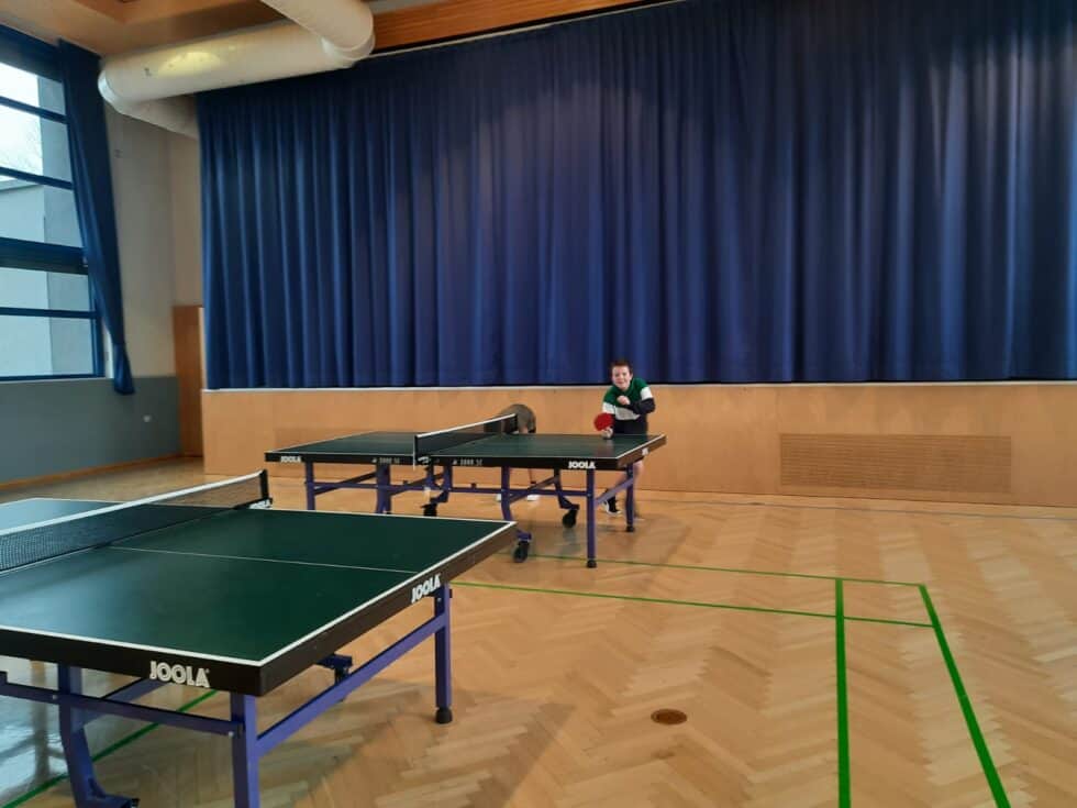 Moderner Turnsaal mit Tischtennistischen, lachendem Spieler und freundlicher Atmosphäre durch natürliche Beleuchtung.